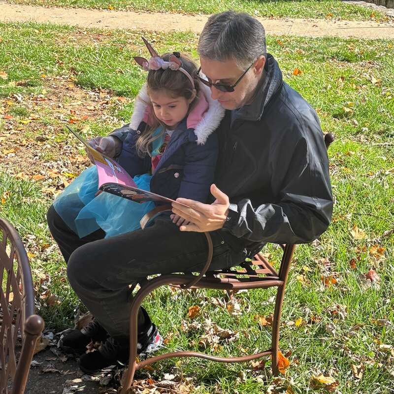 An older man with salt-and-pepper hair reads from a Peanuts Halloween book with his young daughter, who sits in his lap.