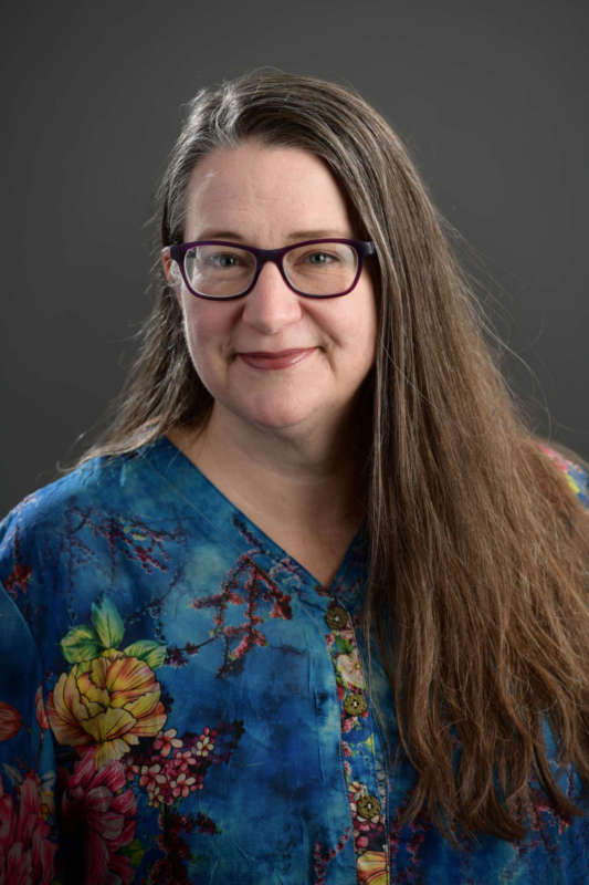 Headshot of Rachael Meads, a white woman with long straight brown hair wearing glasses and a dress with a flowered print.