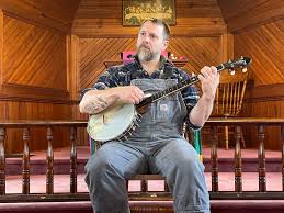 WV musician Ben Townsend wearing overalls sits in a chair playing banjo 