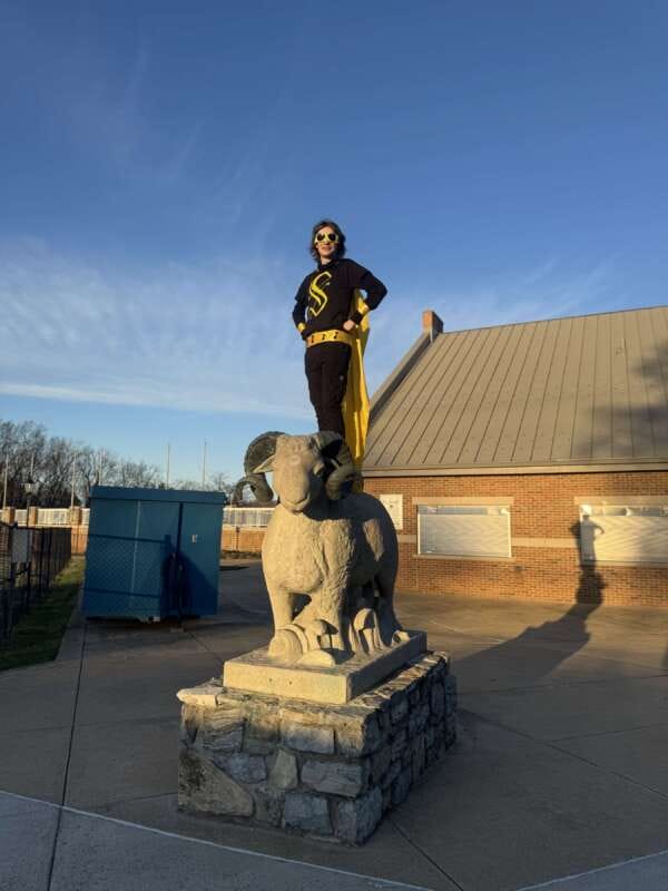 Fraternity member dressed in superhero constume stands on the back of the ϳԹRam Statue at Ram Stadium.