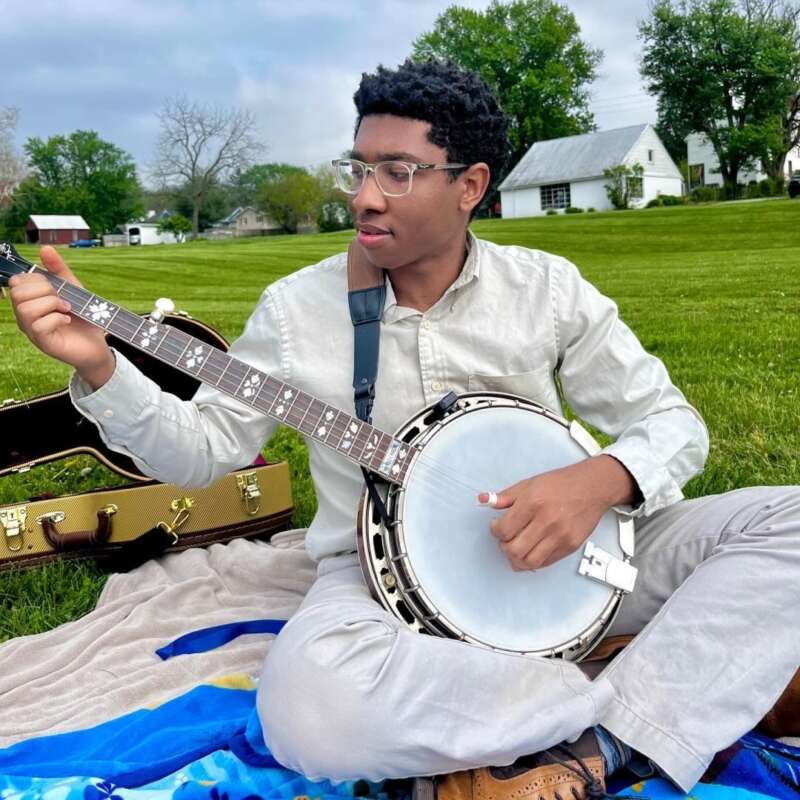 Josh Jimmerson sitting on a blanket in a field playing banjo, with banjo case beside him on a spring day
