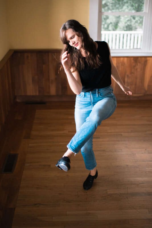 Percussive dancer Becky Hill flatfoots on wooden floor earting jeans and black top. 