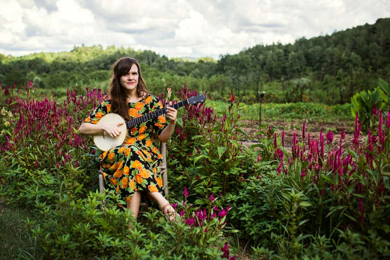 Dancer and musician Becky Hill sits in a chair in a field of purple flowers playing a banjo. Photo by Cory Marie Podielski