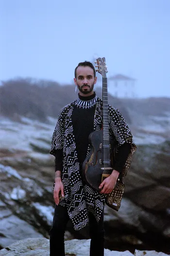 Jake Blount holds a guitar while standing on a rocky ocean shoreline.