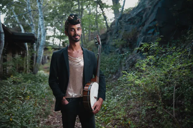 African American folk artist Jake Blount wearing head wrap stands in a forest holding his banjo