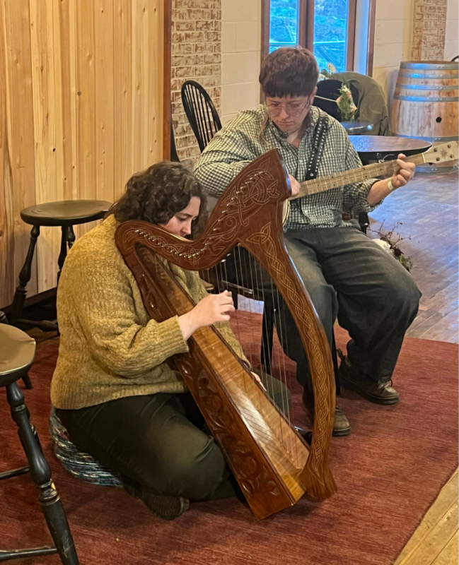 Two musicians, one playing a Celtic harp and one playing a banjo are seated playing on a rug. They are Flora Meeker (banjo) and Fern Poling (Celtic harp)