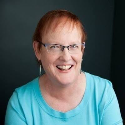 Photo of Dr. Linda Tate, author and scholar. She is a woman with red short hair wearing glasses, handmade earrings, and a turquoise scoop-necked shirt.