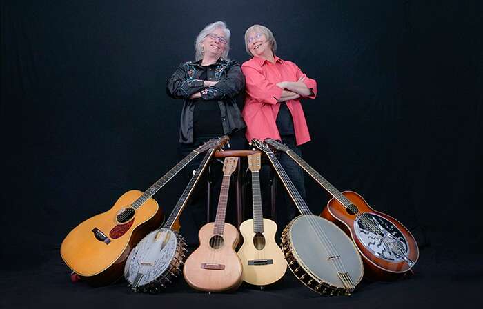 Folk artists Cathy Fink and Marcy Marxer stand shoulder to shoulder surrounded by a variety of instruments including guitars, banjo, mandolin, and resonater guitar.