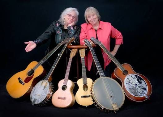 Photo of two female musicians (Cathy Fink and Marcy Marxer) standing side by side surrounded by 6 instruments including two banjos, three guitars, and a steel guitar. 